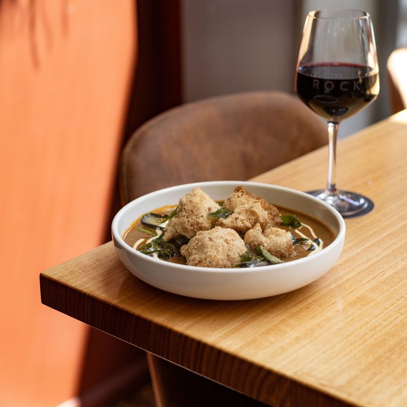 A bowl of curry topped with crackling beside a glass of red wine in a The Rock glass