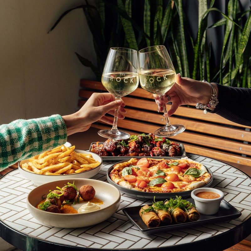 Two glasses of white wine clinking over pizza, wings, chips, arancini and spring rolls